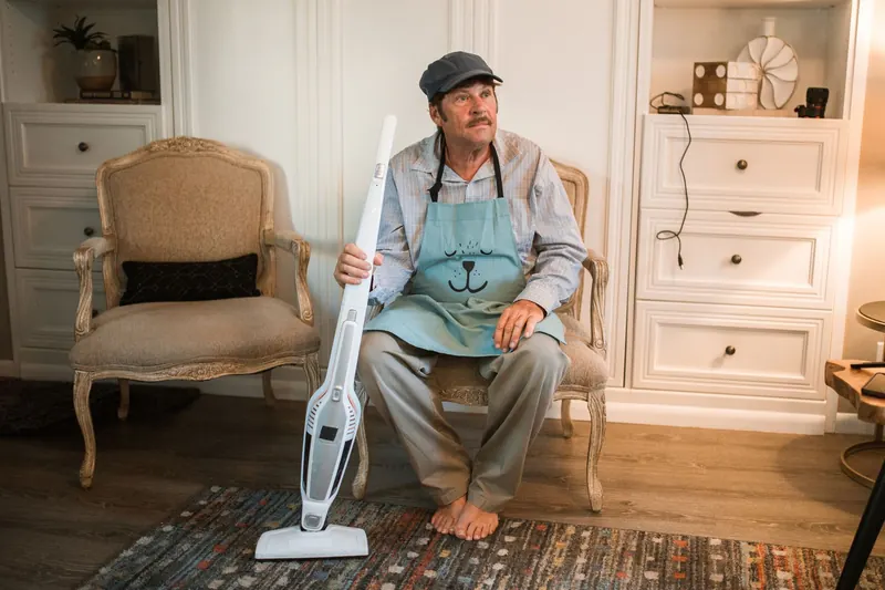 An adult man in a cap and apron holding a vacuum cleaner, sitting on an ornate chair in a cozy living room